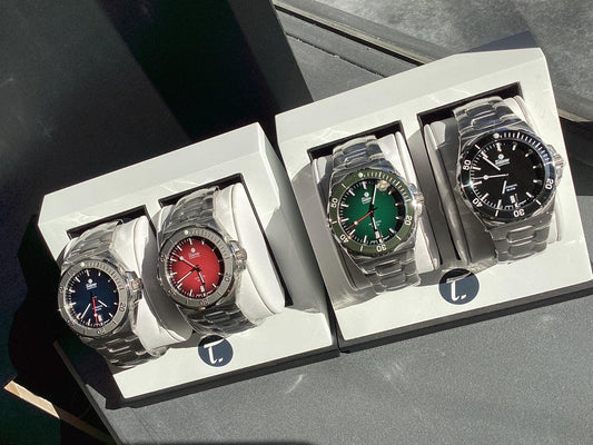 Four Contévanou mechanical wristwatches with red, green, blue, and black dials in display cases with metal bands