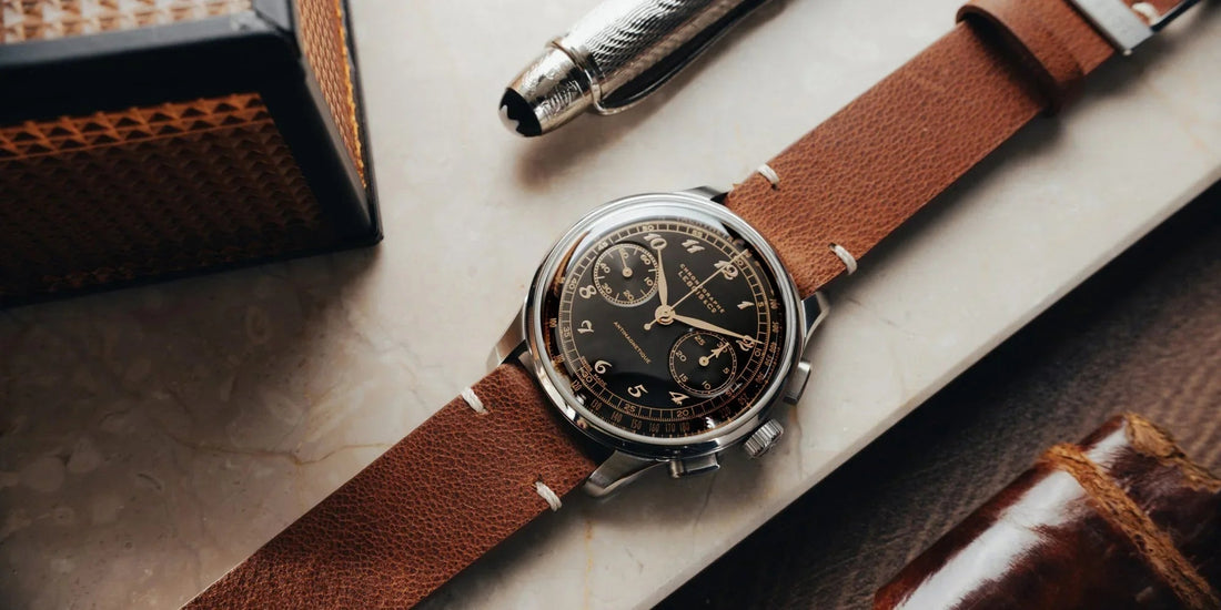 Vintage-style wristwatch with black chronograph dial and brown leather strap on marble surface with luxury pen and leather items